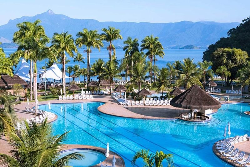 Resort all-inclusive em Angra dos Reis com piscina e vista para o mar, rodeado por palmeiras e montanhas ao fundo.