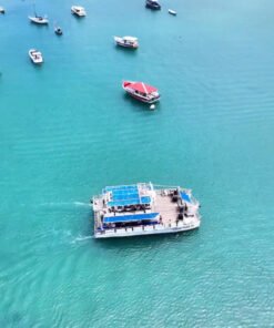 Catamarã navegando em águas cristalinas de Búzios, cercado por barcos menores e com vista para a costa.